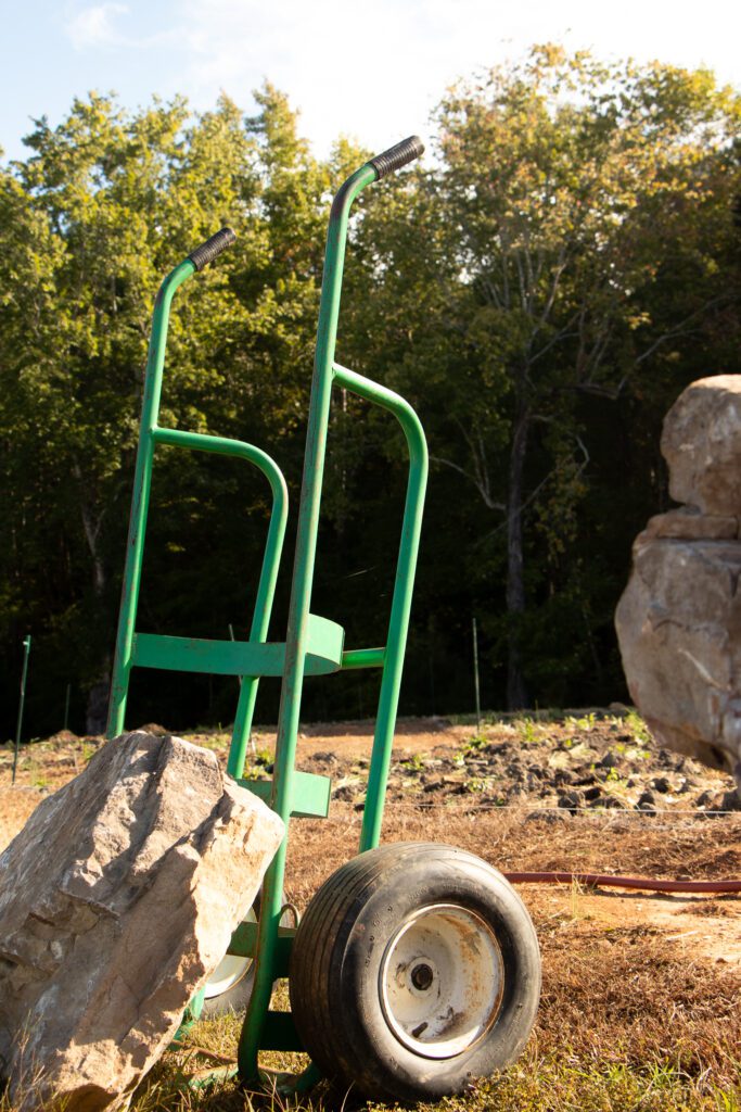 Green metal structure with wheels on rocky ground near trees.